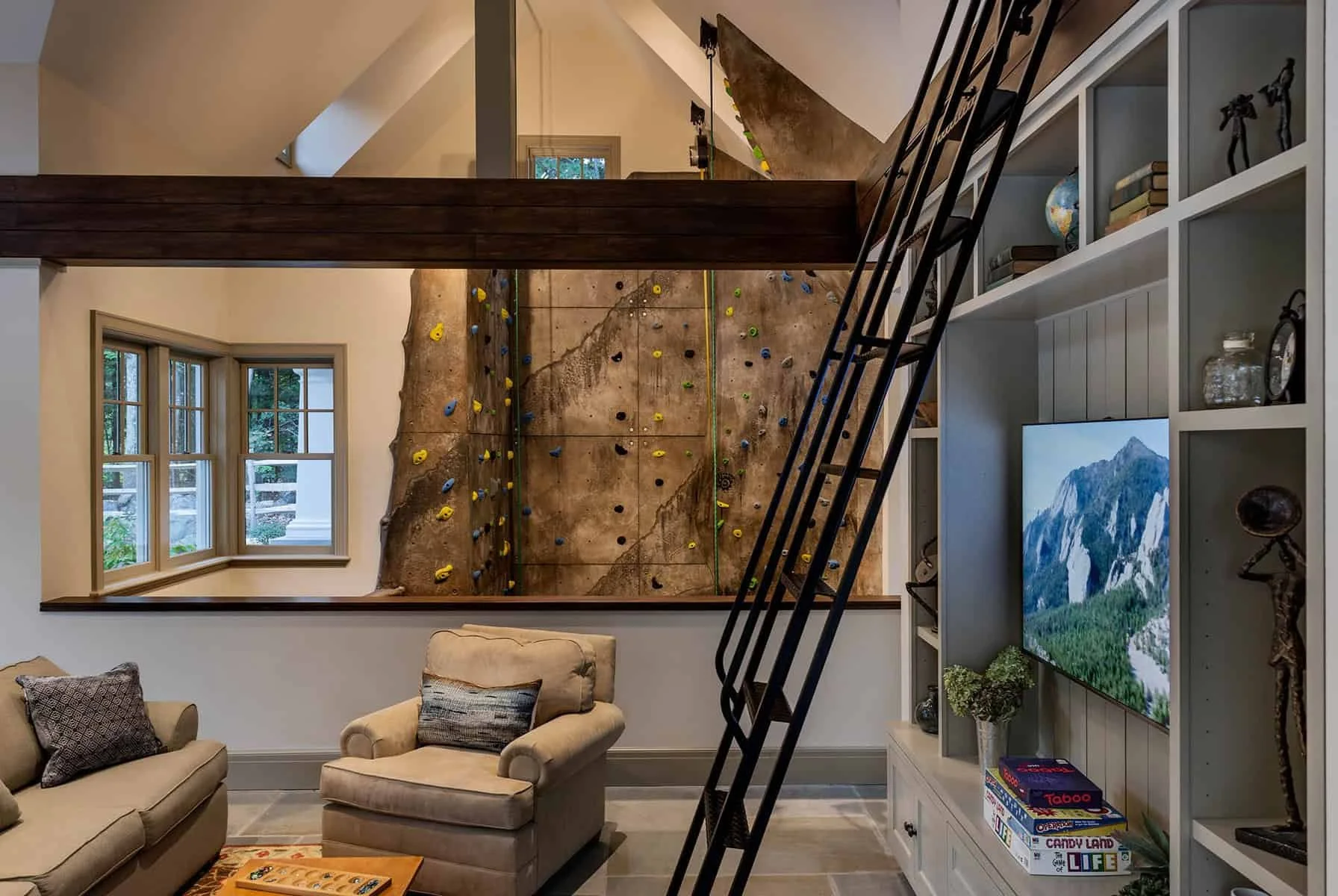 Living room with beige sofas, a plaid pillow, light-colored hardwood floor, a window with multiple panes, a large mountain landscape TV, white built-in shelves with decor and board games, and a rock climbing wall on the back wall.