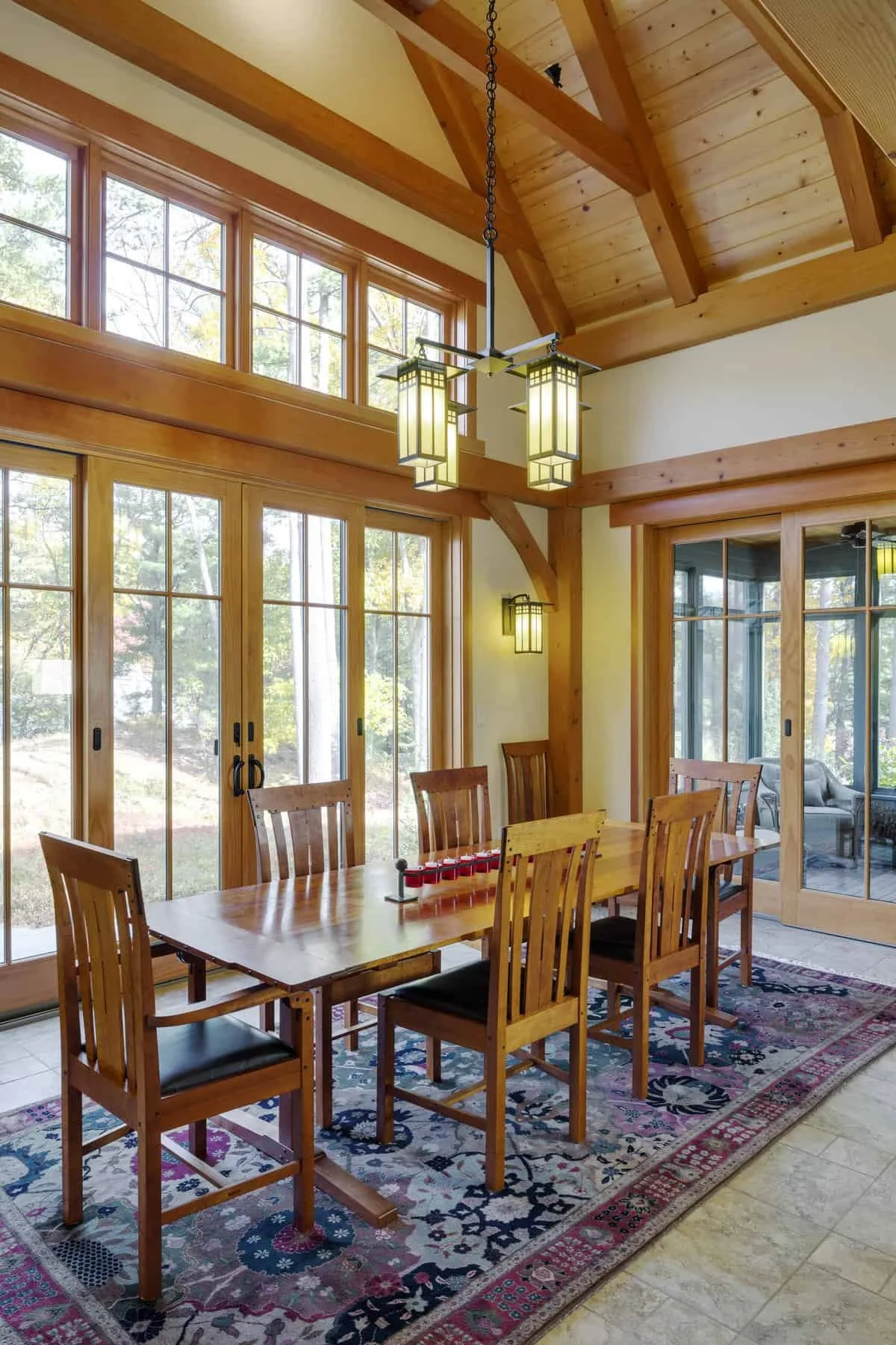A dining room with large windows and wooden beams, featuring a wooden table with eight matching chairs, a colorful area rug, pendant lights, and sliding glass doors leading to an outdoor area with trees and a sitting area visible inside.
