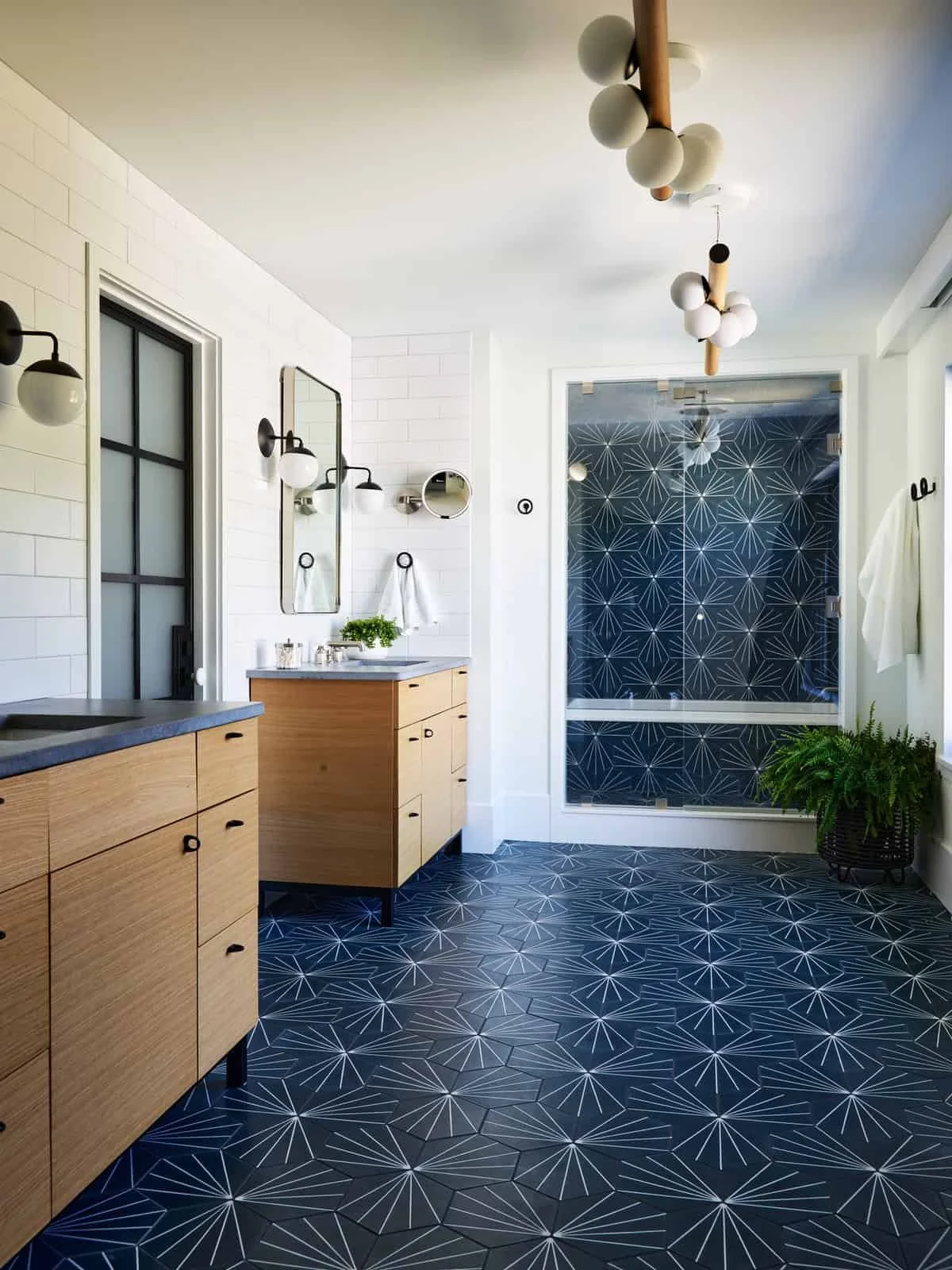 Modern bathroom with white brick walls, wooden vanity cabinets, black and white patterned floor and shower tiles, black wall-mounted light fixtures, large mirror, and a circular mirror. There are plants and towels as decor.