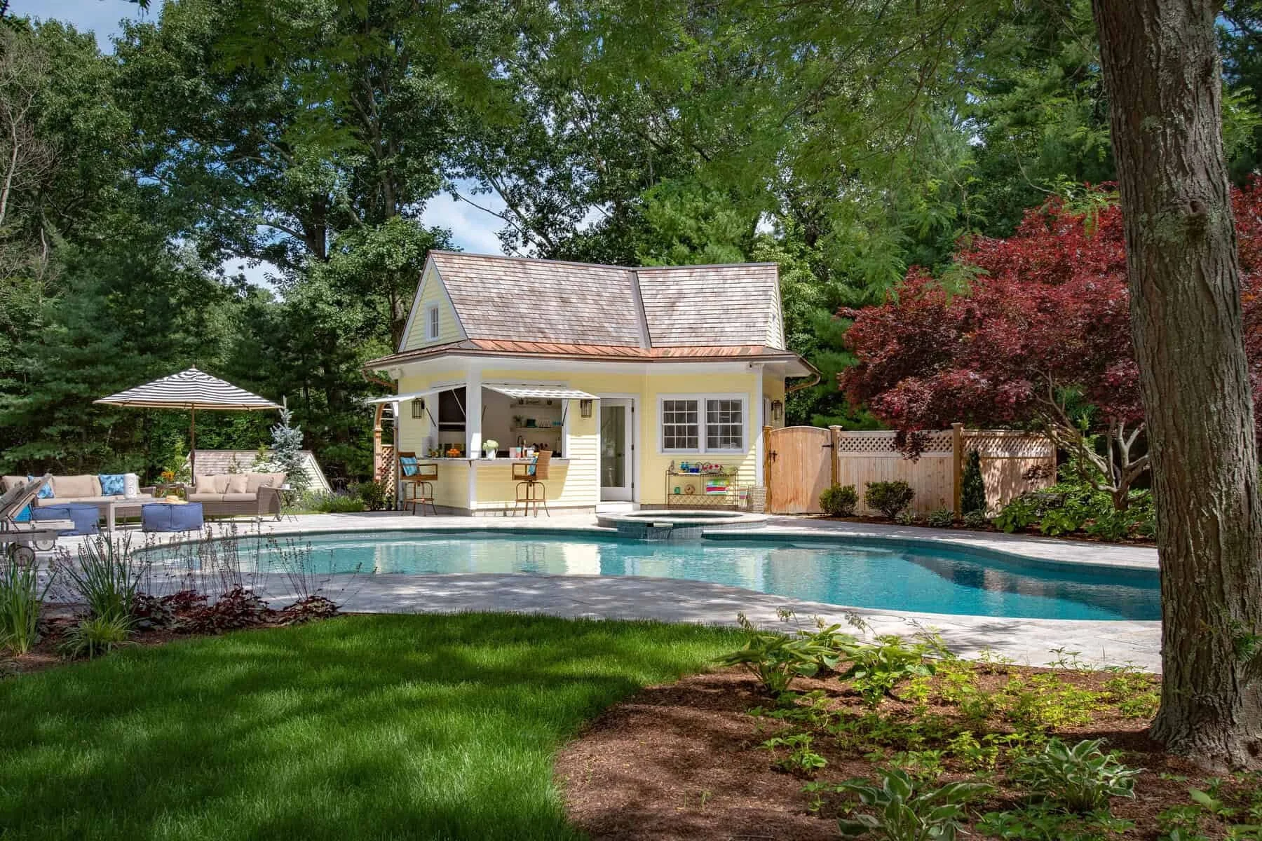A backyard with a swimming pool, outdoor seating with cushions and an umbrella, a small yellow pool house with a bar area, surrounded by trees and shrubs.