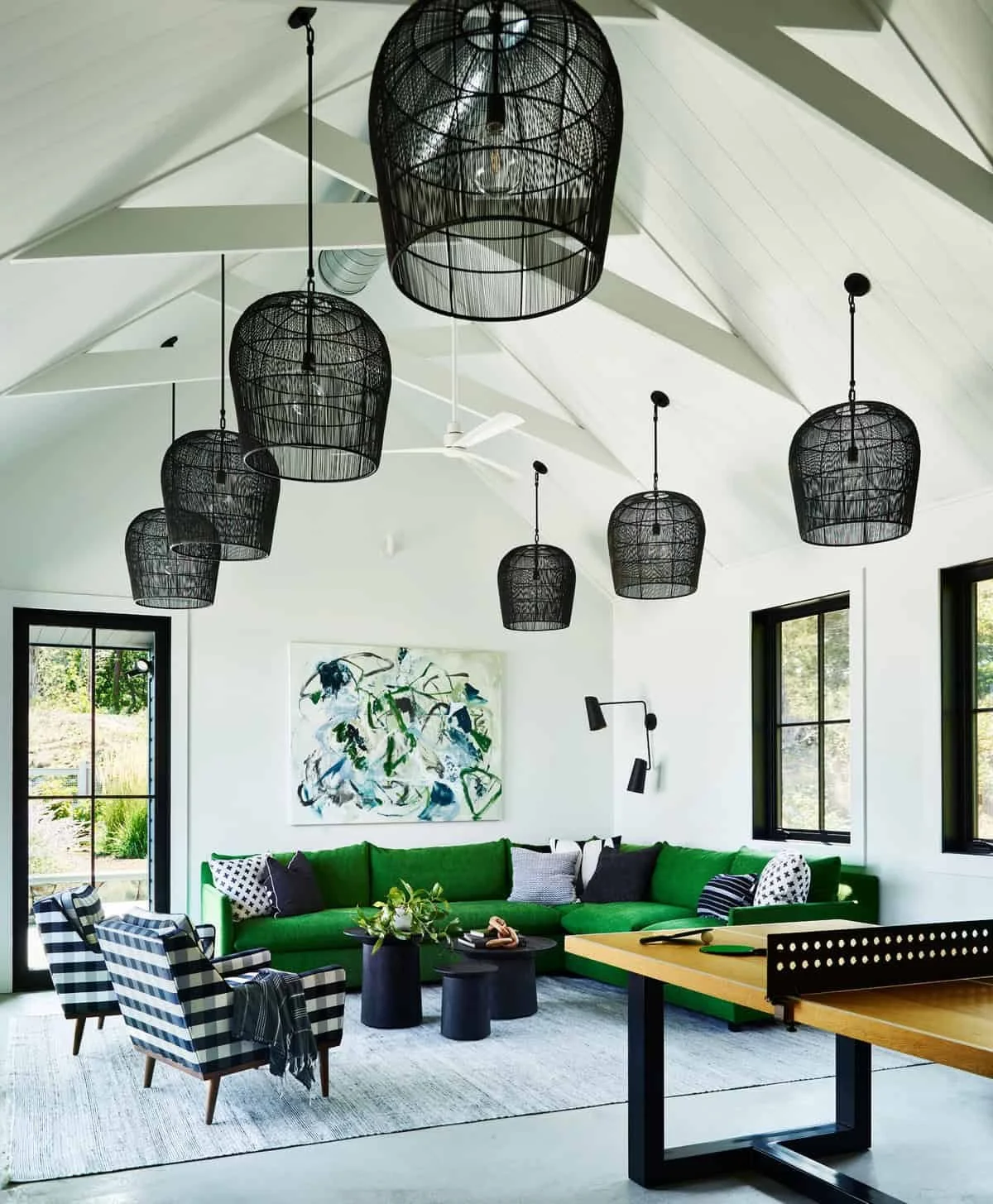 Living room with white walls, black window frames, green couch, black and white patterned chairs, wall art, black pendant lights, and a ping pong table.
