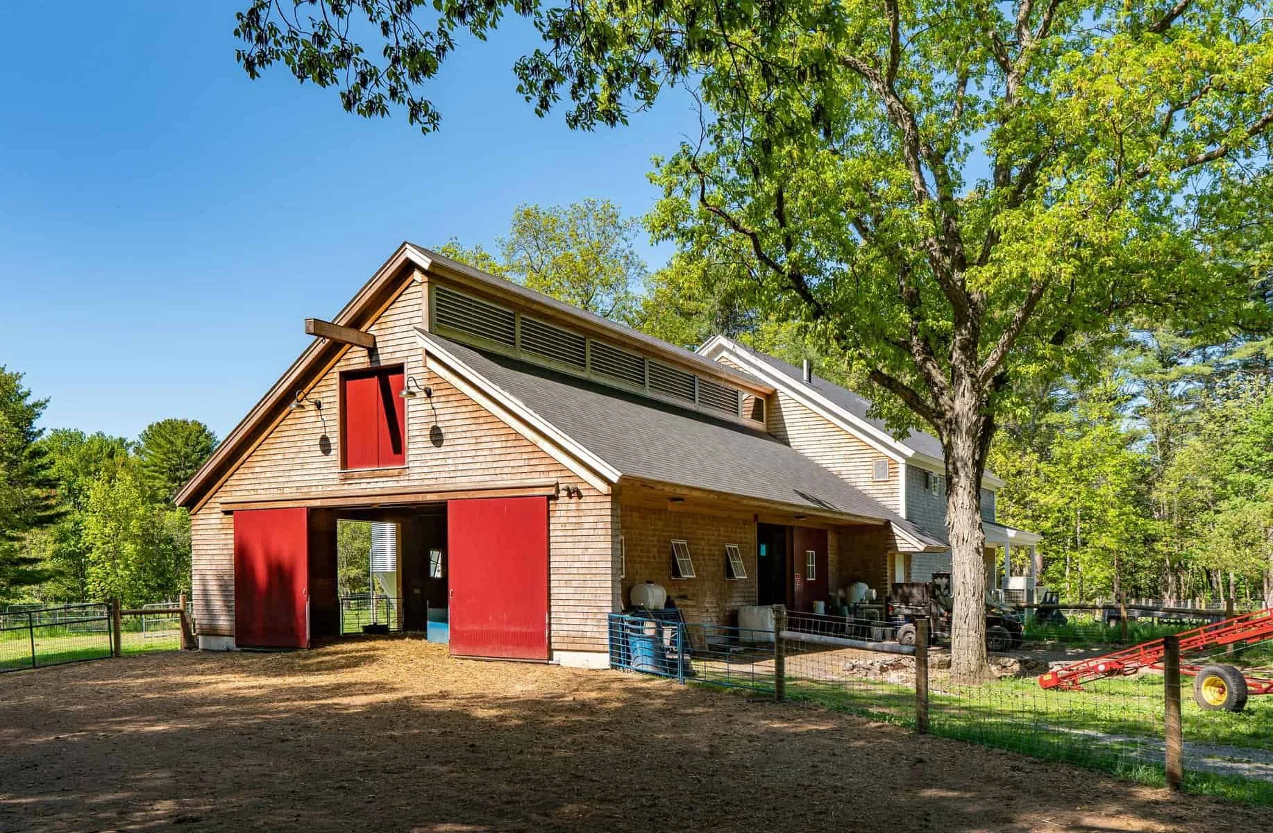A modern barn with red sliding doors, wooden siding, and a sloped roof, surrounded by green trees and a fenced outdoor area.