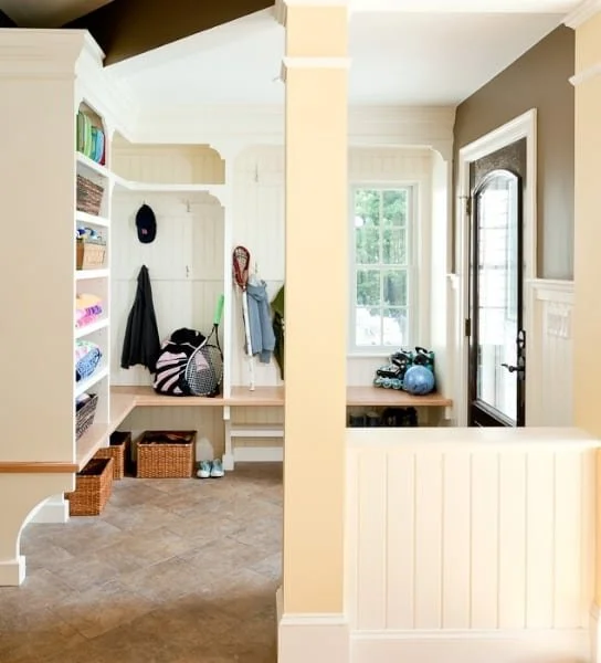 Entryway with white built-in shelves, hooks, baskets, and coat rack, leading to a room with a window, a bench, and a door.