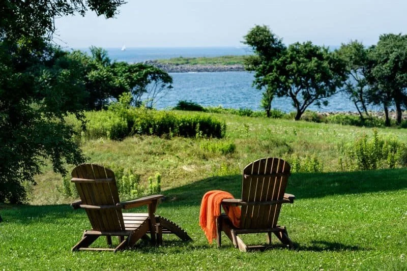 Two wooden Adirondack chairs on a grassy lawn facing a lake with trees and sailboats in the distance.