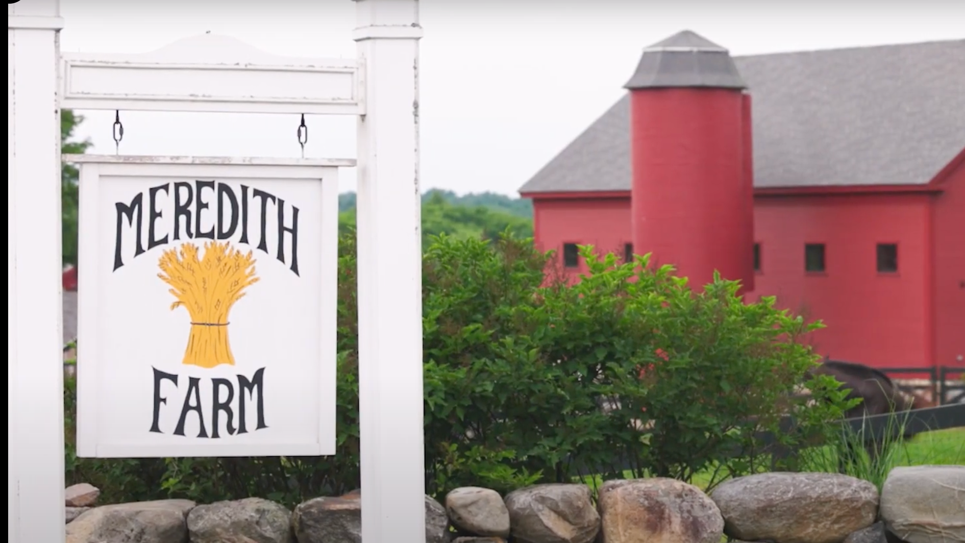 A red barn called Meredith Farm with a gray roof and small windows, surrounded by greenery and rocks.