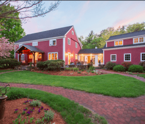 A large red farmhouse-style house with white trim, surrounded by lush green grass and plants, under a pink and blue sky at sunset.
