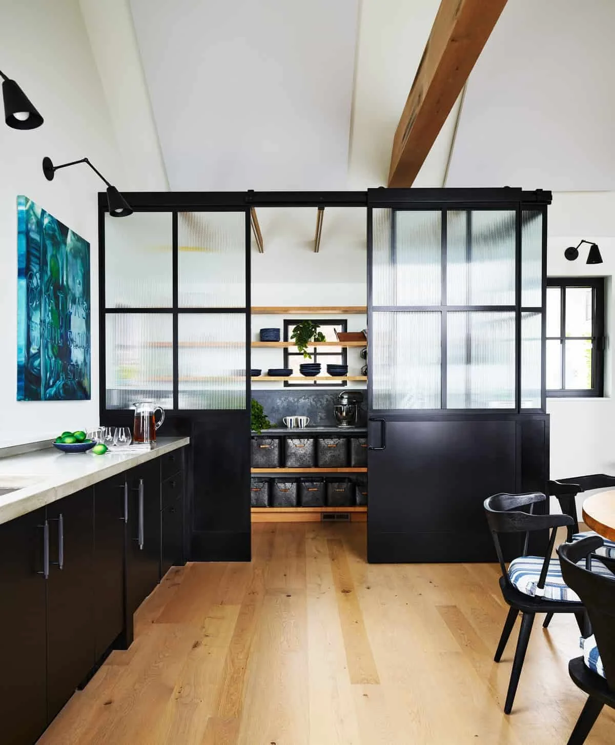 Modern kitchen with black cabinets, wooden shelves, a textured black counter, and a black sliding door with textured glass panels. Open shelving with bowls and a plant, and a kitchen island or breakfast bar on the left. Wooden flooring and a dining t