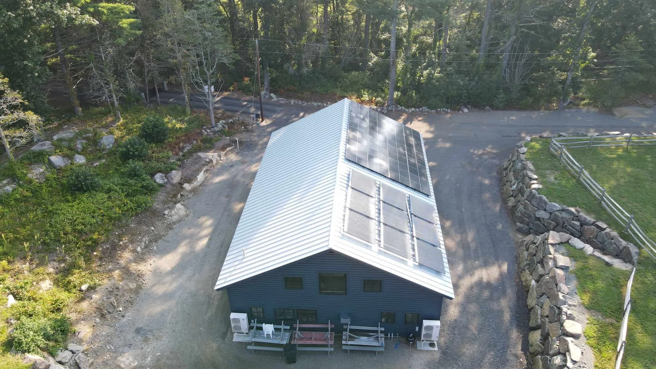 An aerial view of a house with solar panels on its roof, surrounded by trees, a gravel driveway, and a stone-lined yard.