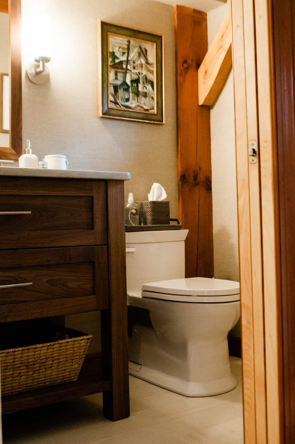 A small bathroom with a wooden vanity, a white toilet, some toiletries on the vanity, a painting on the wall, and a tissue box on top of the toilet tank.