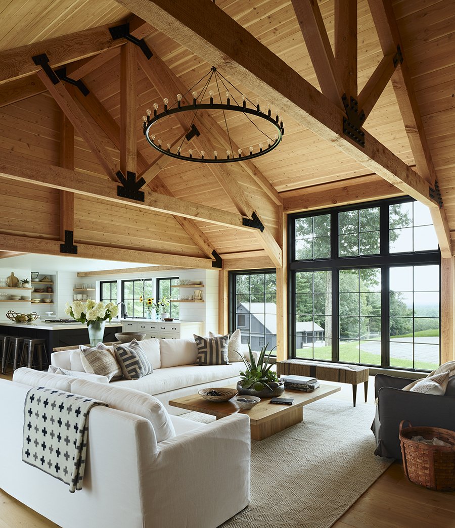 Living room with custom high wood vaulted ceiling, large black window, white sofa with patterned pillows, wooden coffee table with plants and books, and black armchair, overlooking lush green landscape.
