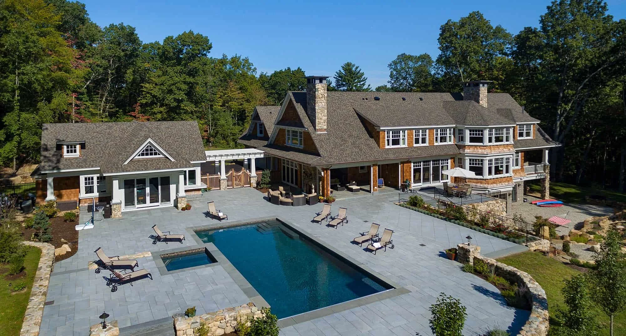 A large house with multiple levels and a gray shingle roof, surrounded by trees. The backyard features a rectangular swimming pool with lounge chairs and an outdoor seating area with umbrellas, all on a paved patio.