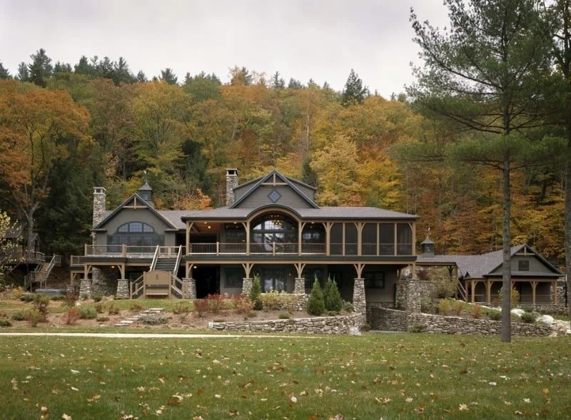 Large house with multiple gabled roofs, stone columns, and a wrap-around porch, set against a backdrop of autumn-colored trees.
