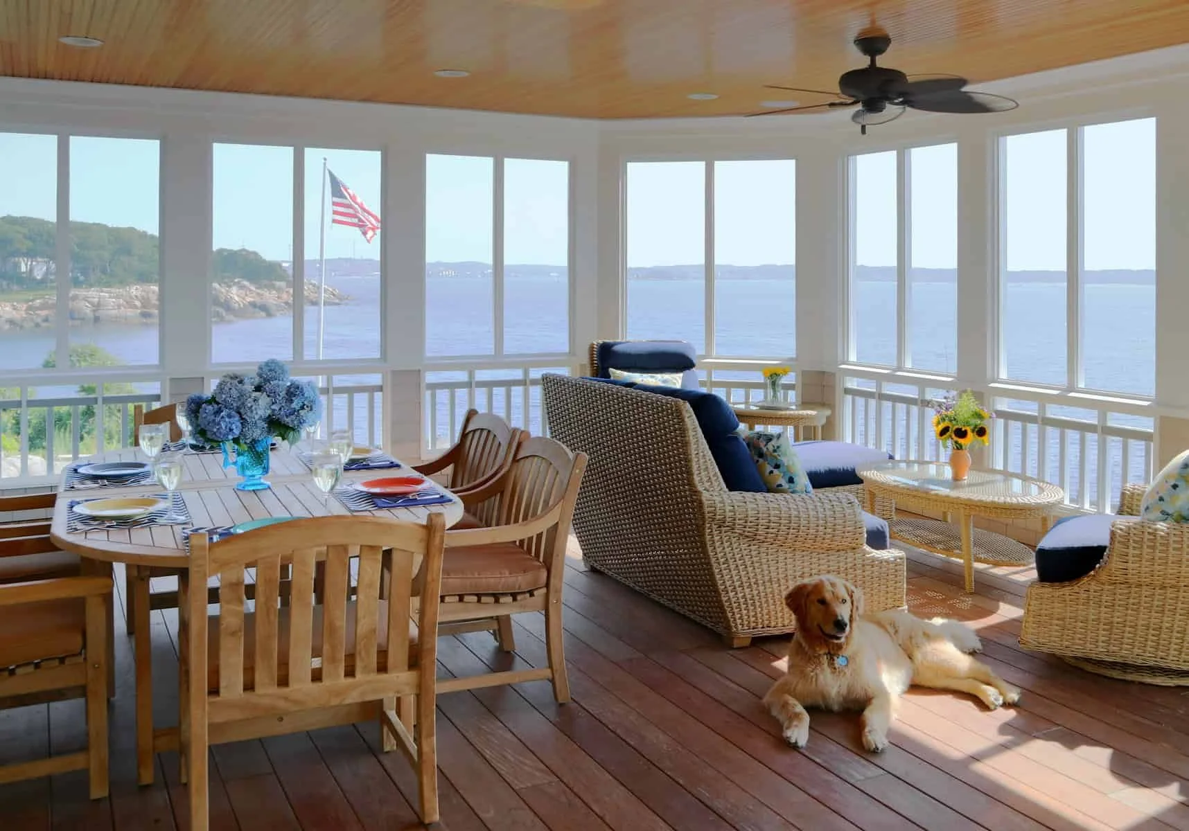Sunlit enclosed porch overlooking water with American flag, wooden dining table set for a meal, wicker and wooden chairs with cushions, a vase of blue flowers, a dog lying on the wooden floor, and wicker and upholstered seating with colorful pillows.