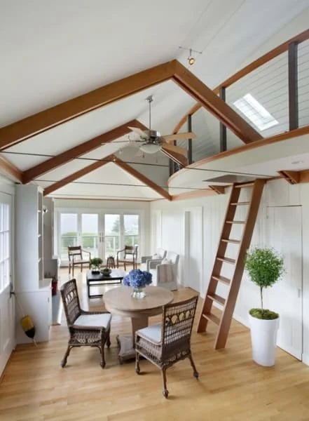 Bright living room with a lofted ceiling, wooden beams, a ceiling fan, a round dining table with four chairs, large windows, a potted plant, and a small lounge area near the windows.