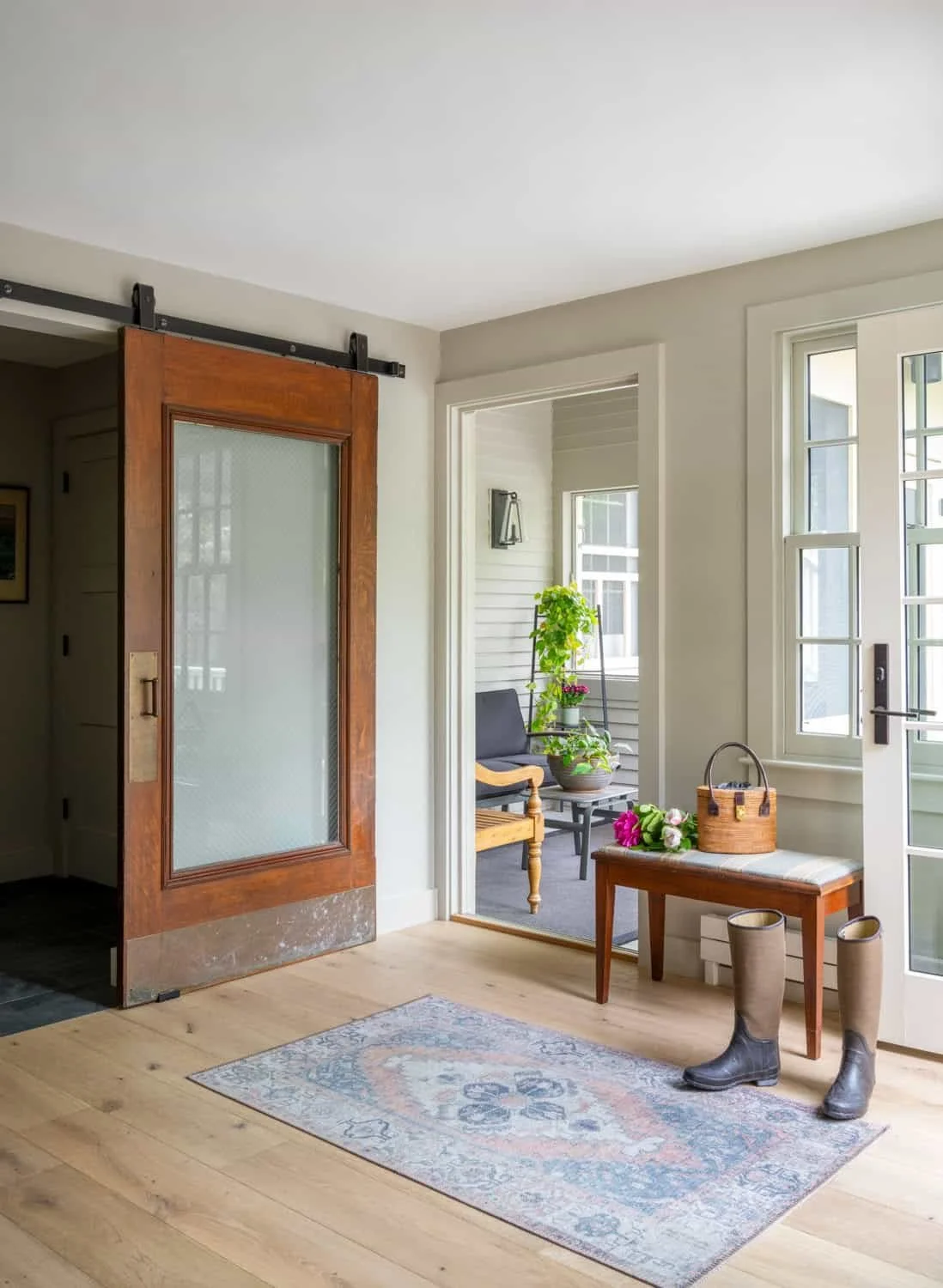 Home entryway with wooden floor, rug, rain boots, a wooden bench with flower bunch, a basket, and a small table with plants on porch.