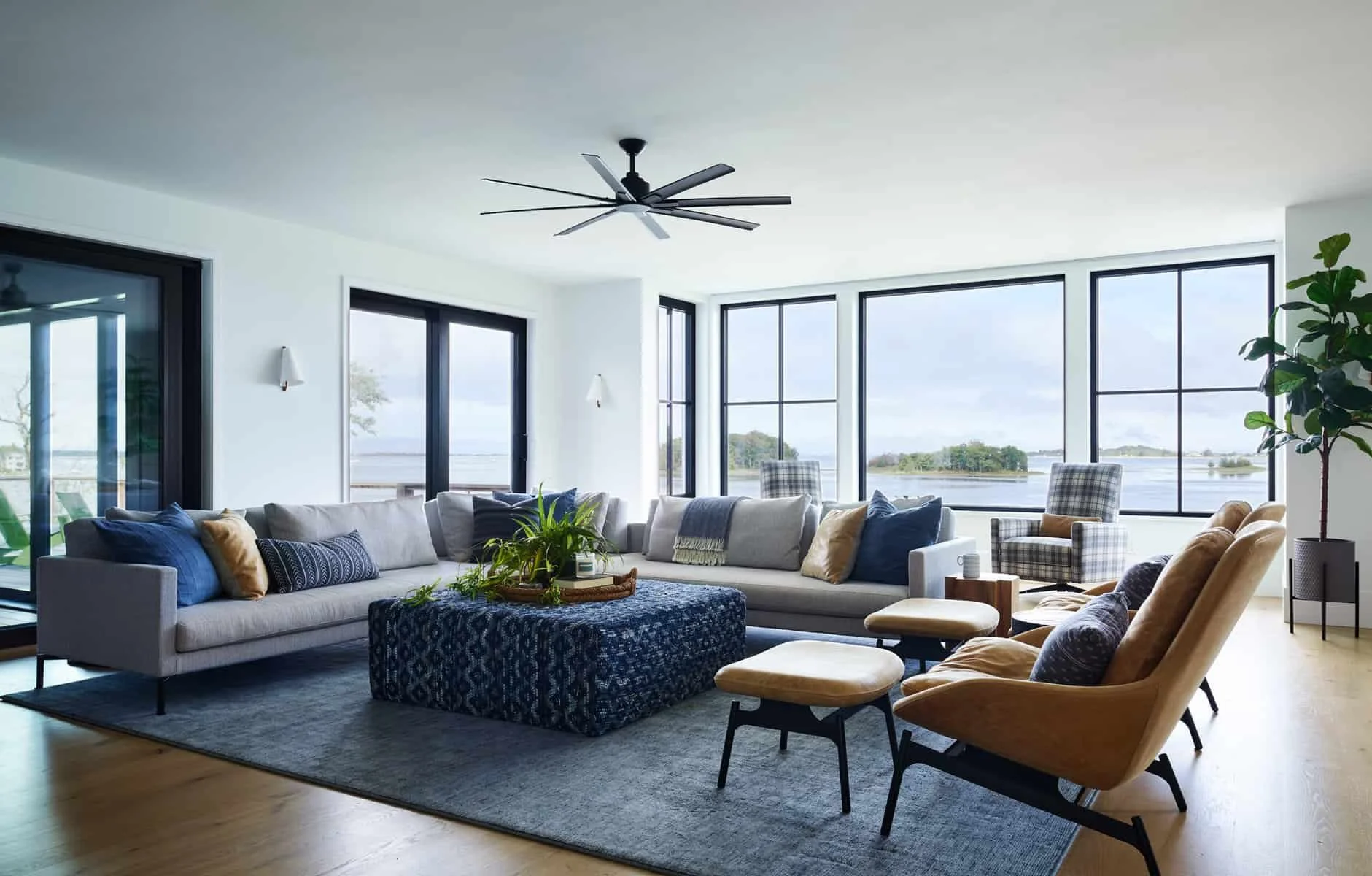 Bright living room with large windows overlooking water, featuring gray sectional sofa with blue and beige pillows, patterned armchairs, a dark blue ottoman, and indoor plants.