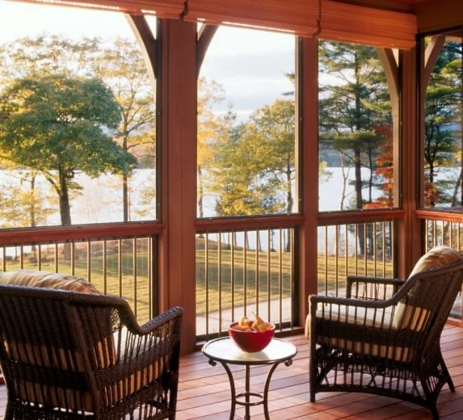 A screened porch with wicker chairs and a small table holding a red bowl of snacks, overlooking a lakeside scene with trees and water during sunset.