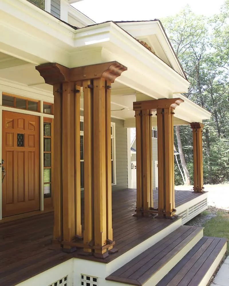 Front porch with four wooden columns supporting the roof of the house, a wooden front door, and steps leading up to the porch, surrounded by trees.