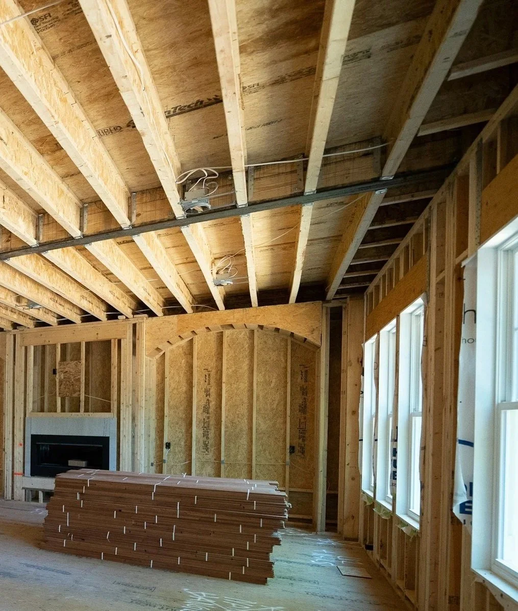 Interior view of a house under construction with exposed wooden framing, drywall and insulation surfaces, and a stack of wooden flooring planks.