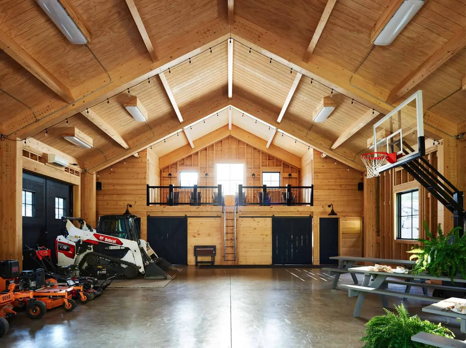 Interior view of a spacious wooden garage or barn with a high vaulted ceiling, string lights, and windows at the back. On the left side, there are farm equipment and a Bobcat skid-steer loader. On the right, a basketball hoop is mounted near a window