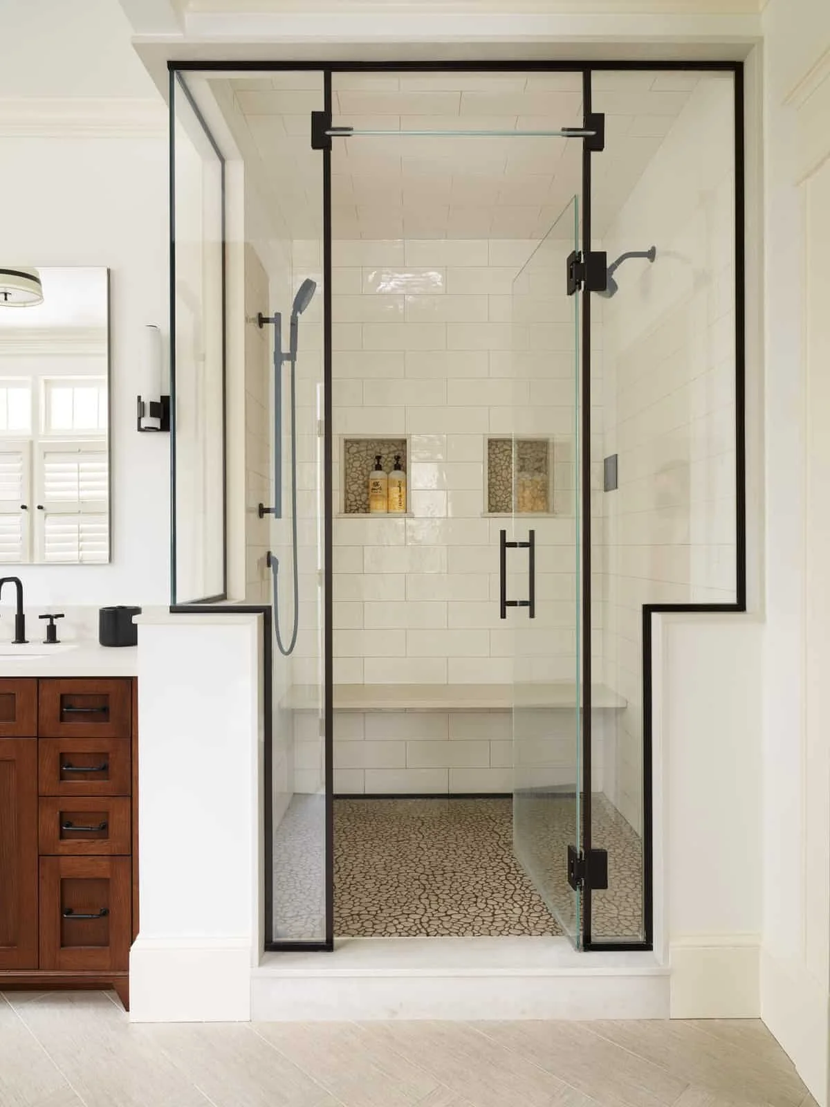 Modern glass-walled shower with black framing, built-in niches with toiletries, pebble flooring, white subway tile walls, and a wooden vanity with black fixtures inside a bathroom.