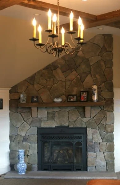 Living room fireplace with a stone surround and a wooden mantel, decorated with framed photos and ornaments, illuminated by a chandelier with lit candles.