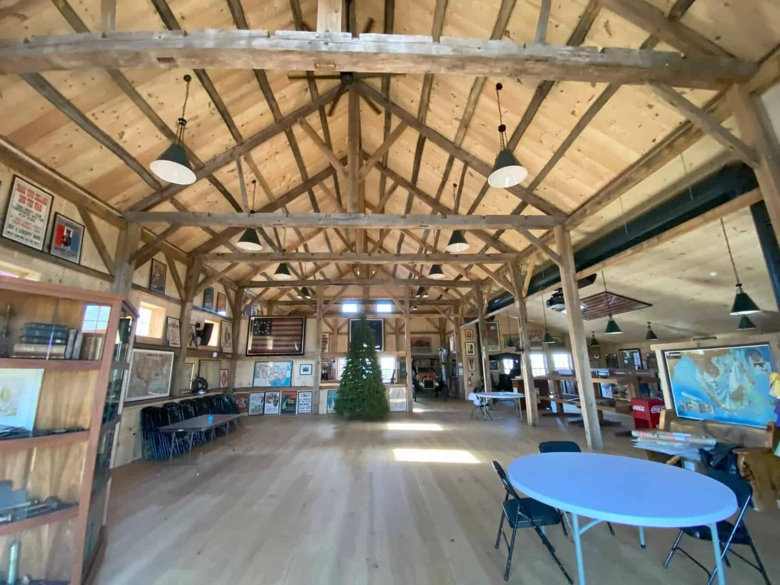 The interior of a wooden exhibition or community hall with a high vaulted ceiling and exposed beams, decorated with framed pictures and American flags, featuring a Christmas tree at the center, black chairs along the walls, a round table with chairs 