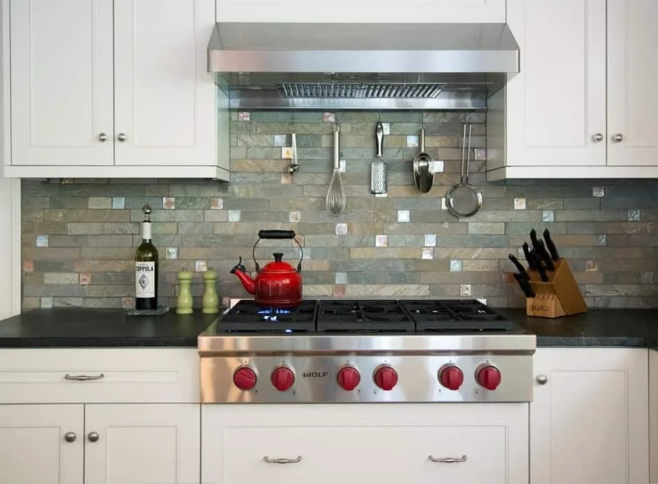 Kitchen stove with four red control knobs, a red kettle on the burner, and kitchen utensils hanging on a backsplash with stone tiles. White cabinets on either side, a knife set in a wooden block, and a bottle of wine with two salt and pepper shakers 