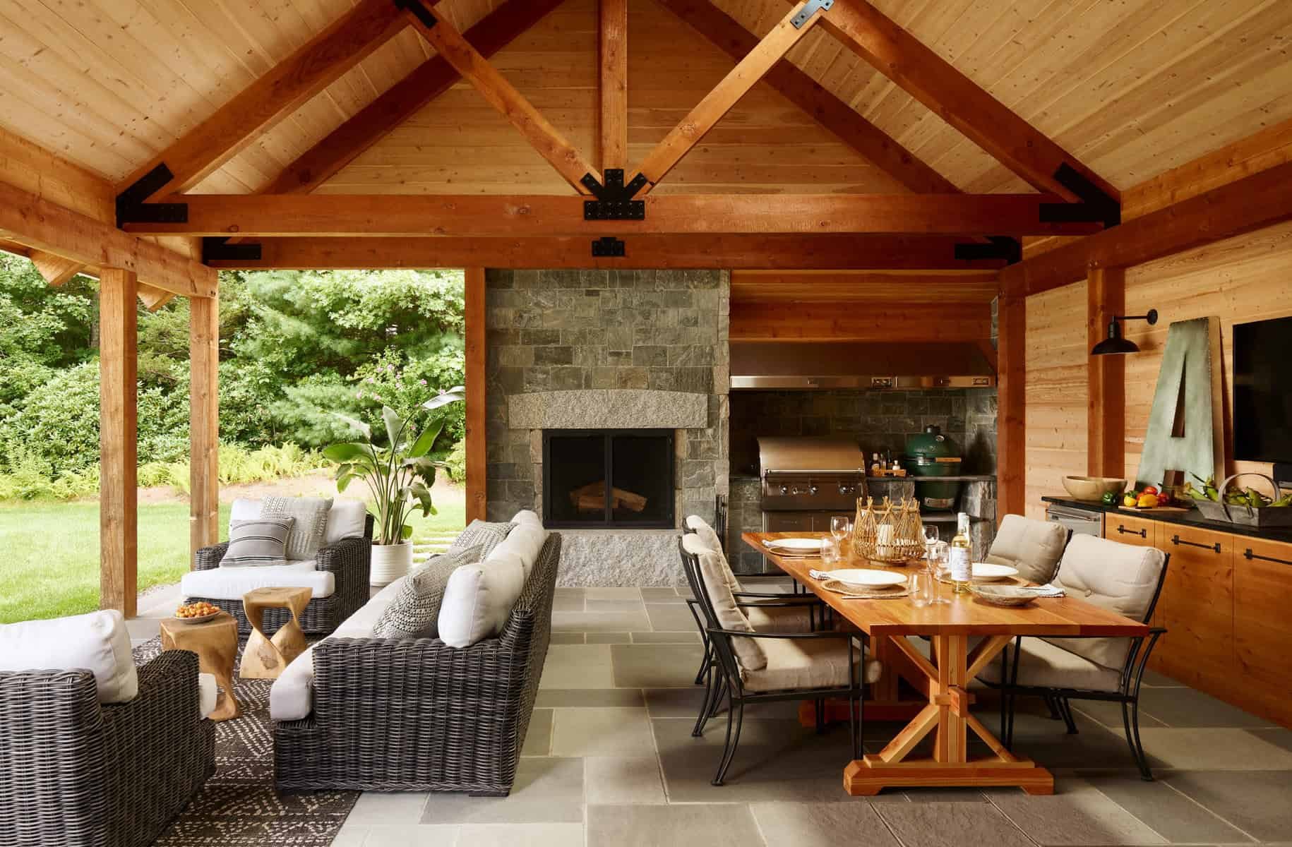Open-air living space with a wooden ceiling and beams, a stone fireplace, seating area with wicker chairs and cushions, a dining table with chairs, and a view of greenery outside.