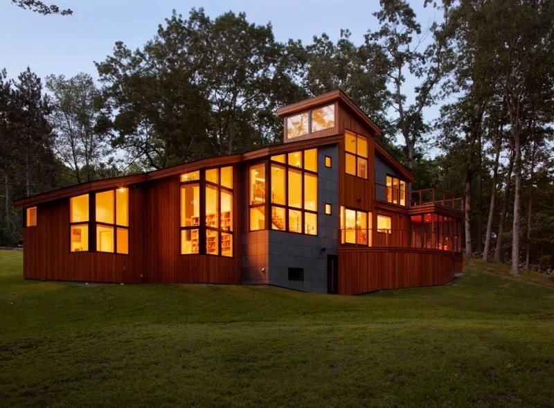 Modern house with large windows showing warm interior lights, surrounded by trees and green grass at dusk.