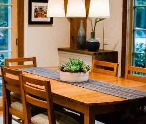 Wood dining table with six matching chairs, a decorative centerpiece, pendant lights, and windows with curtains in a cozy dining room.