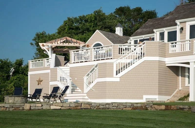 A multi-level beige and white house with a large outdoor deck and staircase, surrounded by a grassy yard and trees in the background.