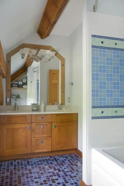 Bathroom with a double sink vanity, large mirror, wooden cabinetry, tiled blue and white shower, and sloped ceiling with wooden beam.