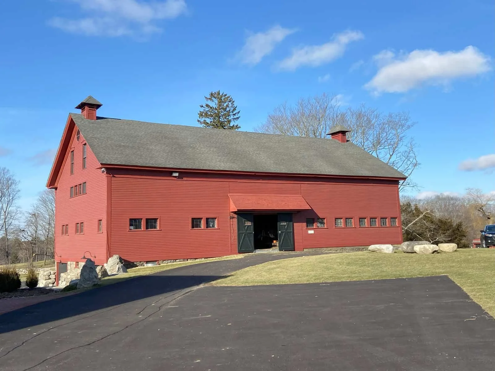 A large red barn with black double doors open, situated on a well-maintained lawn with a paved driveway in front, under a blue sky with scattered clouds and leafless trees in the background.