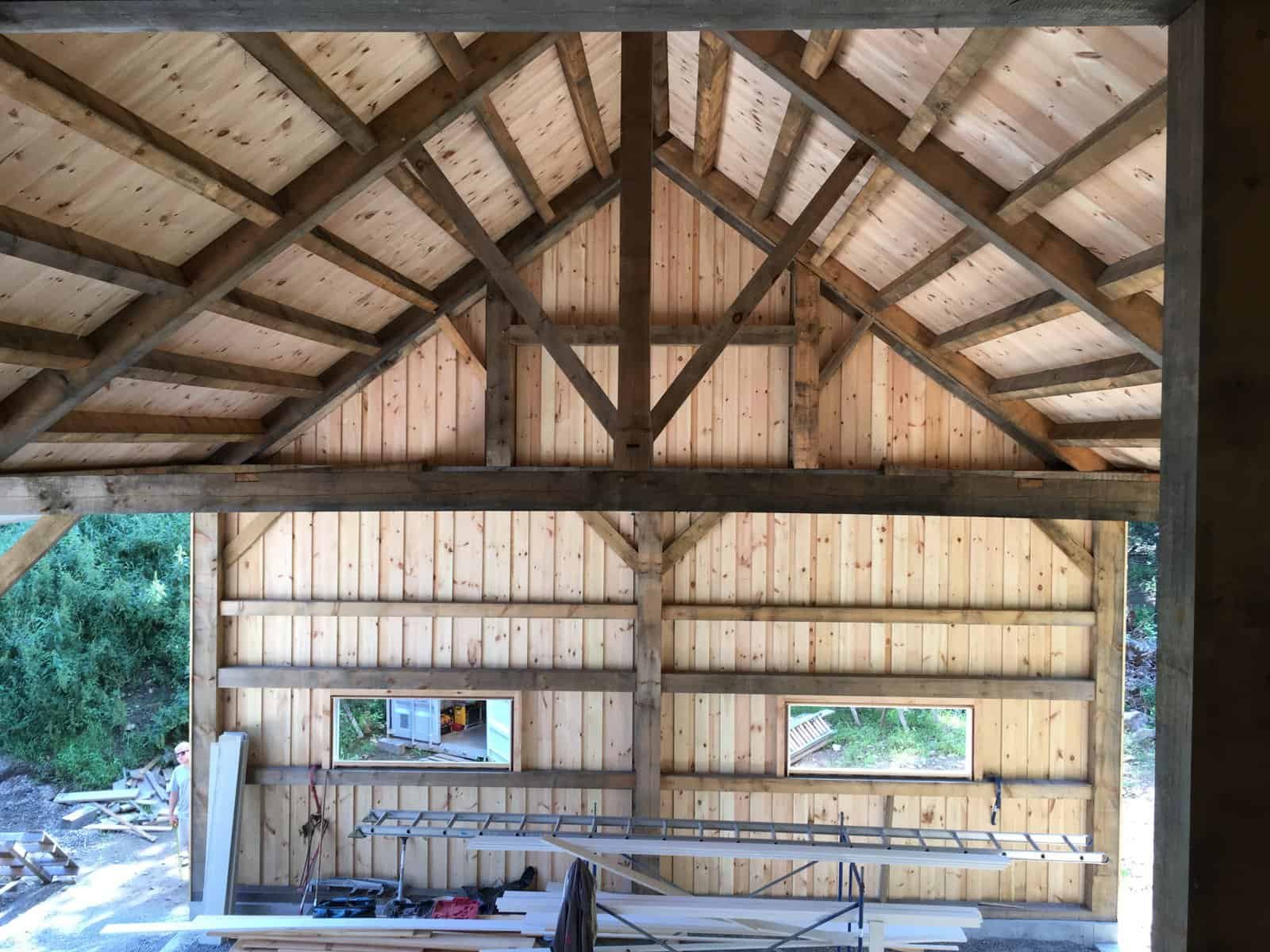 Interior view of a wooden barn under construction, showing the exposed roof trusses, wall framing, and construction materials.