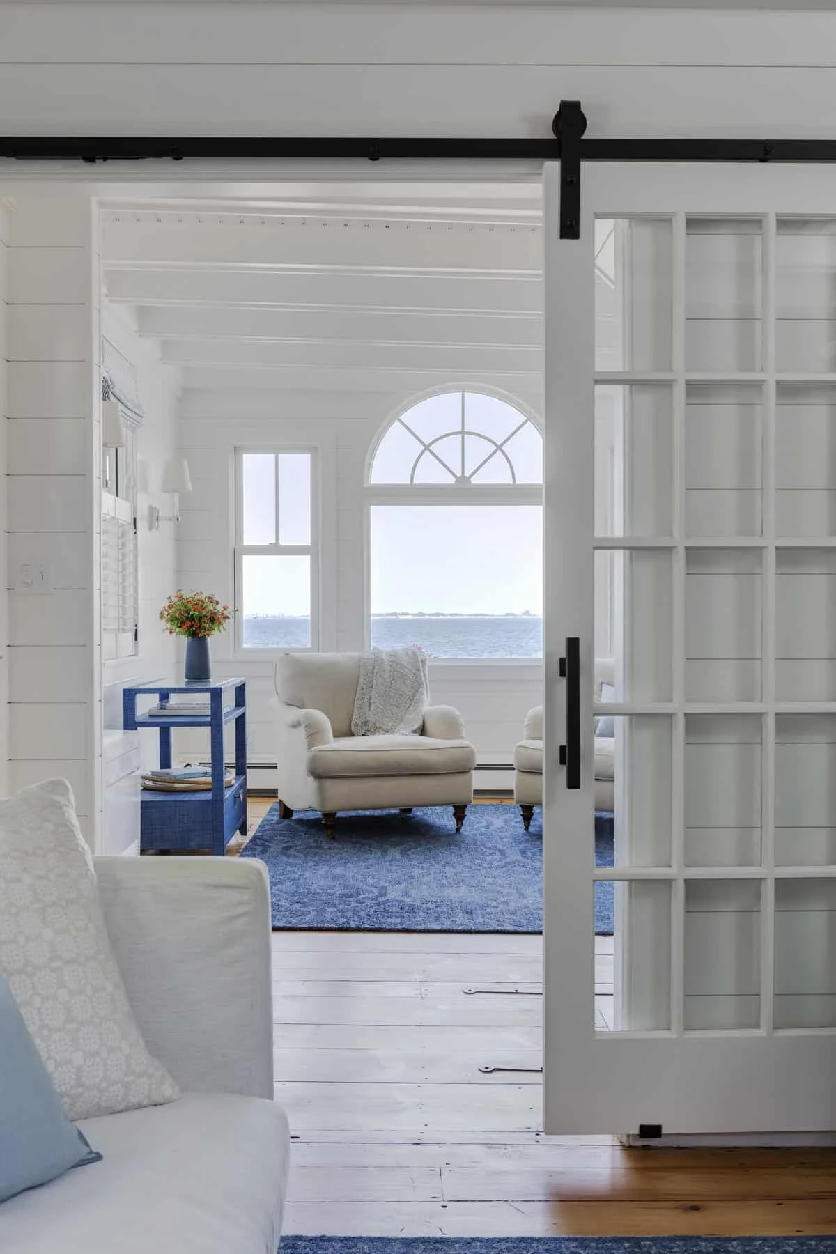 Bright living room with large arched window overlooking water, white walls, beige armchairs, a blue side table with a vase of flowers, and a blue rug.