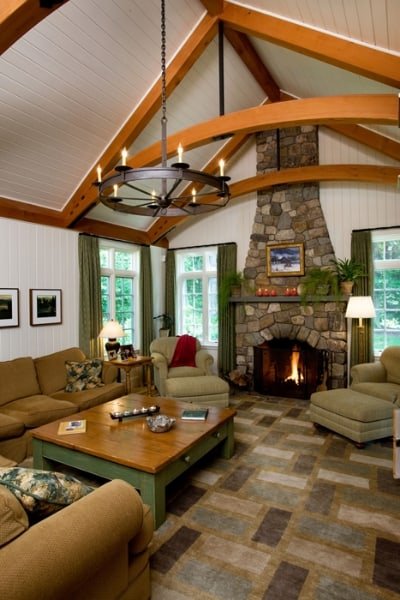 Living room with beige sofas, a stone fireplace, wooden beams on the ceiling, windows with green curtains, and a chandelier.