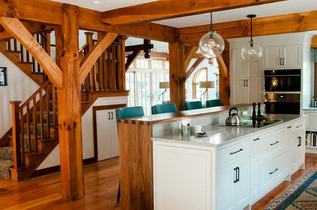 Kitchen with wooden beams and staircase, white cabinetry, a kitchen island with teal chairs, and hanging glass light fixtures.