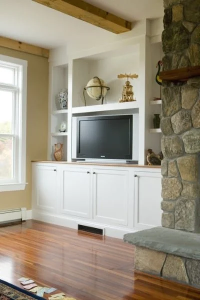 Living room with built-in white cabinet, a flat-screen TV, decorative items on shelves, a stone fireplace, and hardwood flooring.