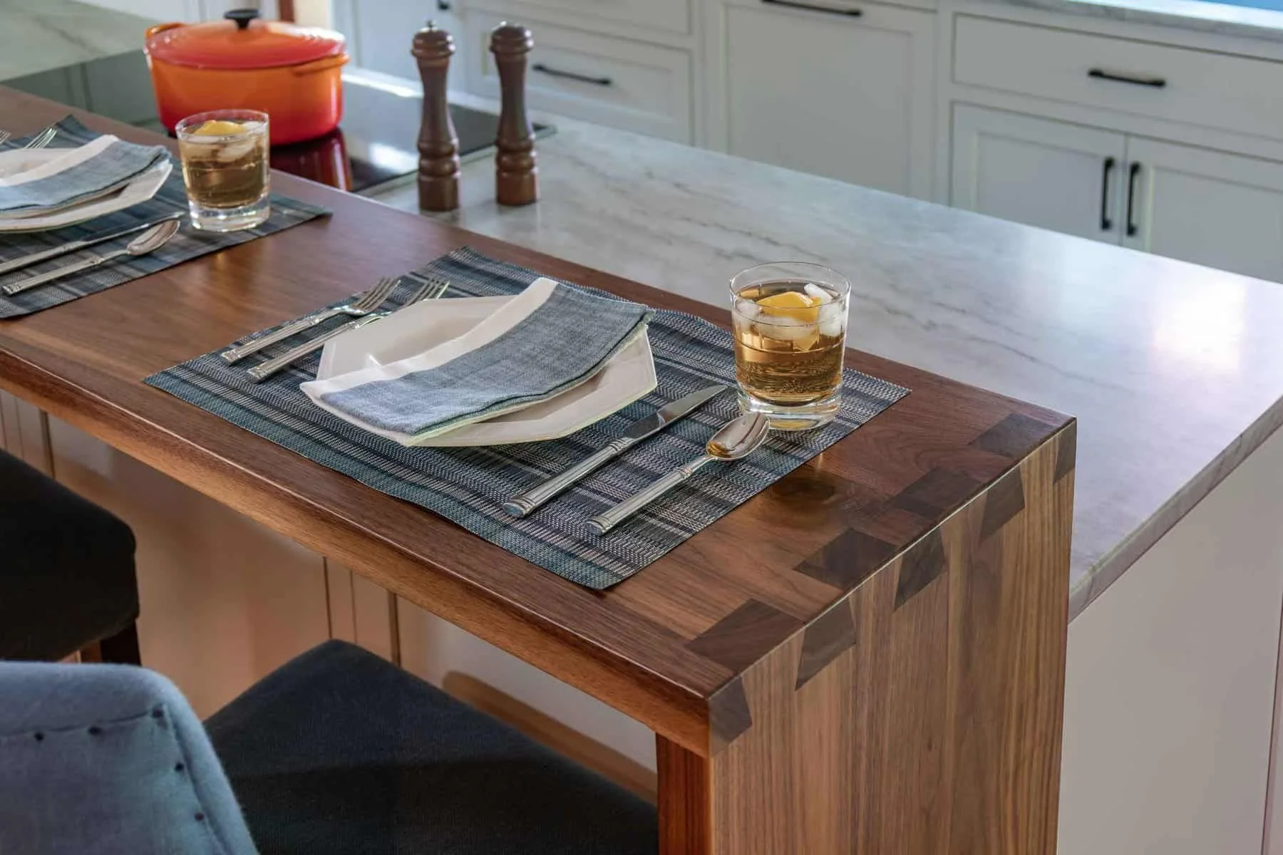 A kitchen dining table set for two with placemats, white plates, cloth napkins, and utensils. Two glasses of iced tea with lemon slices are placed on the table. The table has a wooden surface and a marble countertop in the background. There are salt 