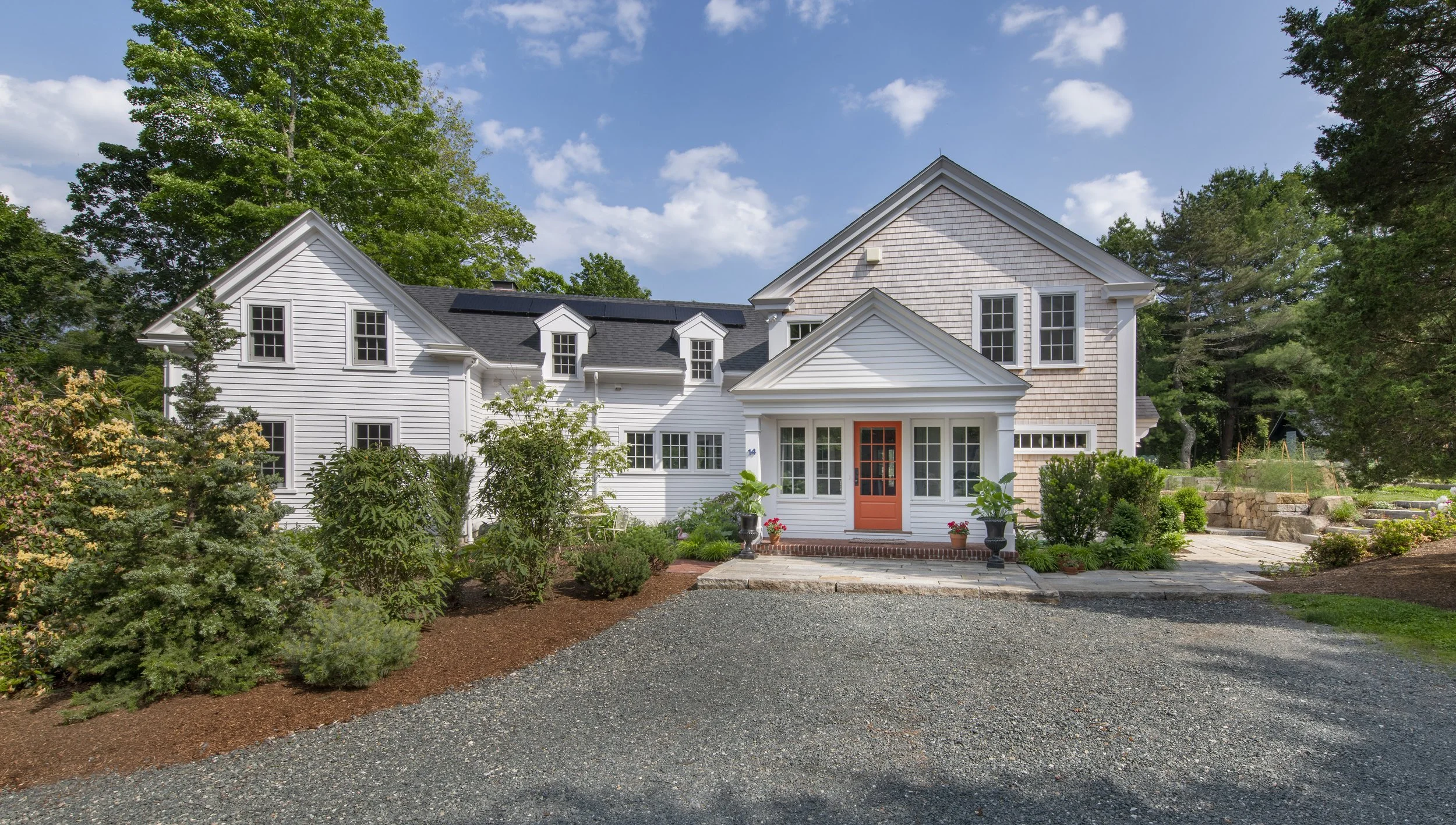 White two-story house with a red front door, black roof, and multiple windows, surrounded by green trees and a garden, under a partly cloudy sky.