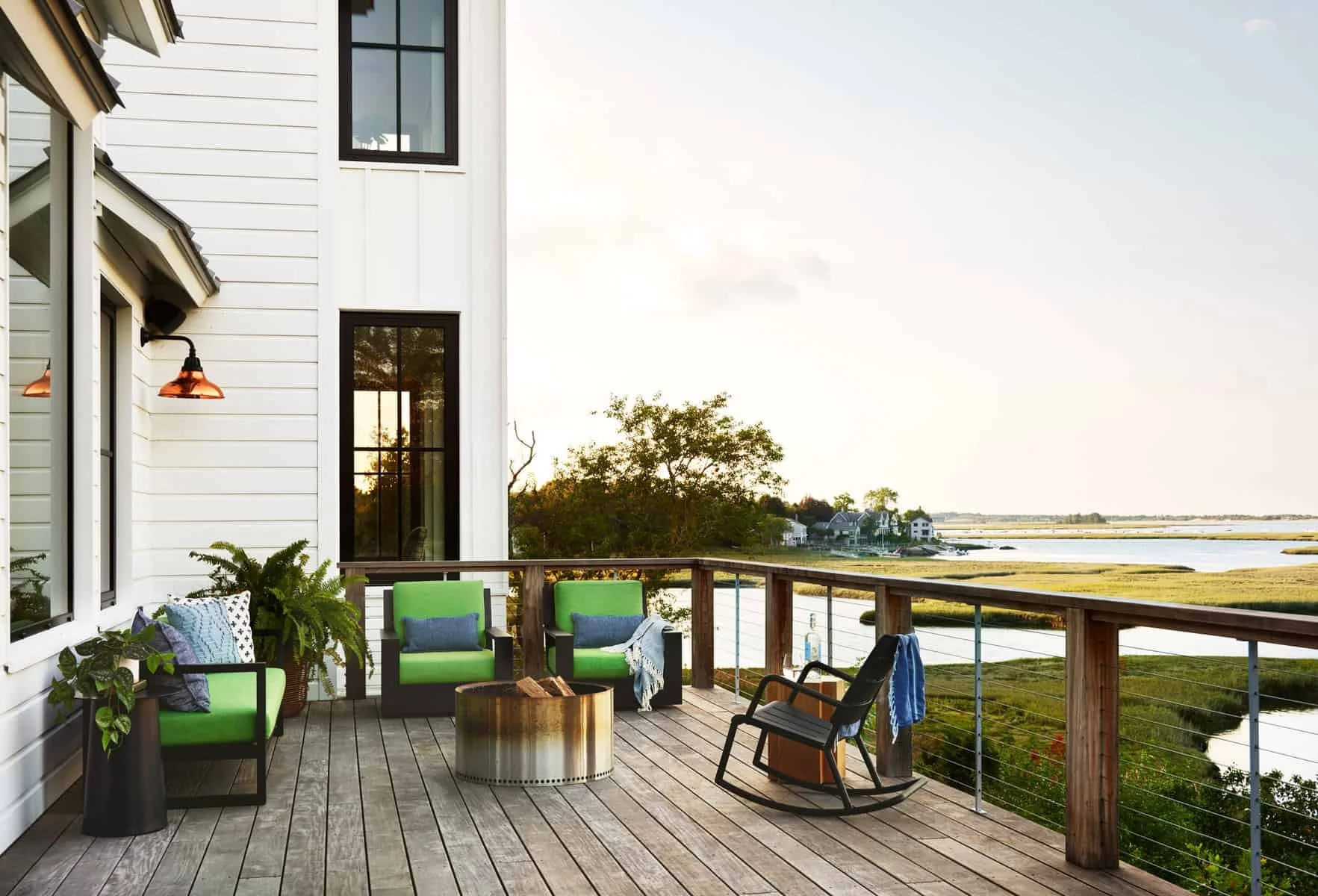A balcony with wooden flooring and a railing overlooking a body of water and green marshland. There are three chairs, two with bright green cushions, a round wooden table, and a black rocking chair. Potted plants and patterned pillows are on the balc