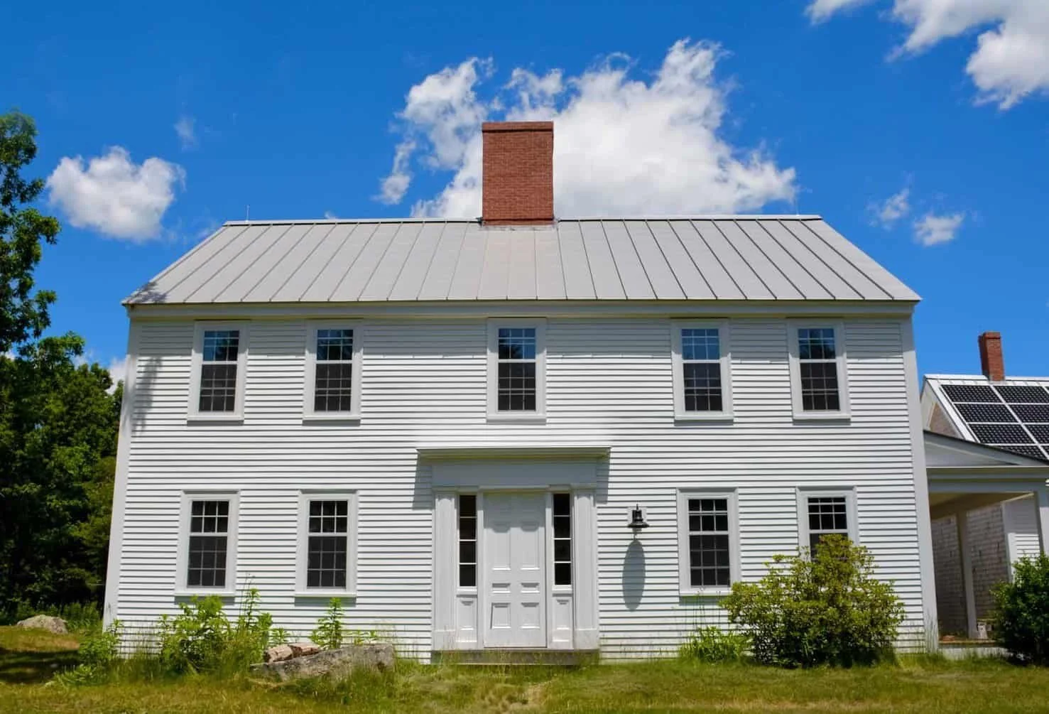 White two-story house with a metal roof and brick chimney under a blue sky with clouds, surrounded by green trees and bushes, with solar panels on a neighboring roof.