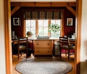 Home office with wooden desk, two chairs, a window with blinds, a potted plant, and framed pictures on dark red walls.