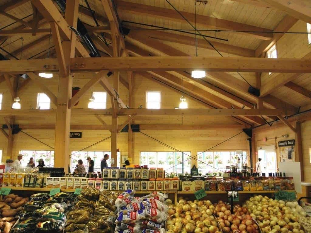 Interior of a grocery store with wooden beams, customers shopping, and fresh produce like potatoes, onions, and packaged goods on display.