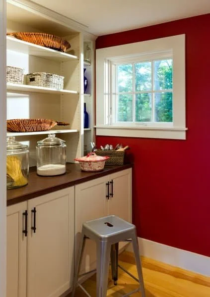 Corner of a kitchen with white cabinets, open shelves with baskets, a red wall, and a window with greenery outside.