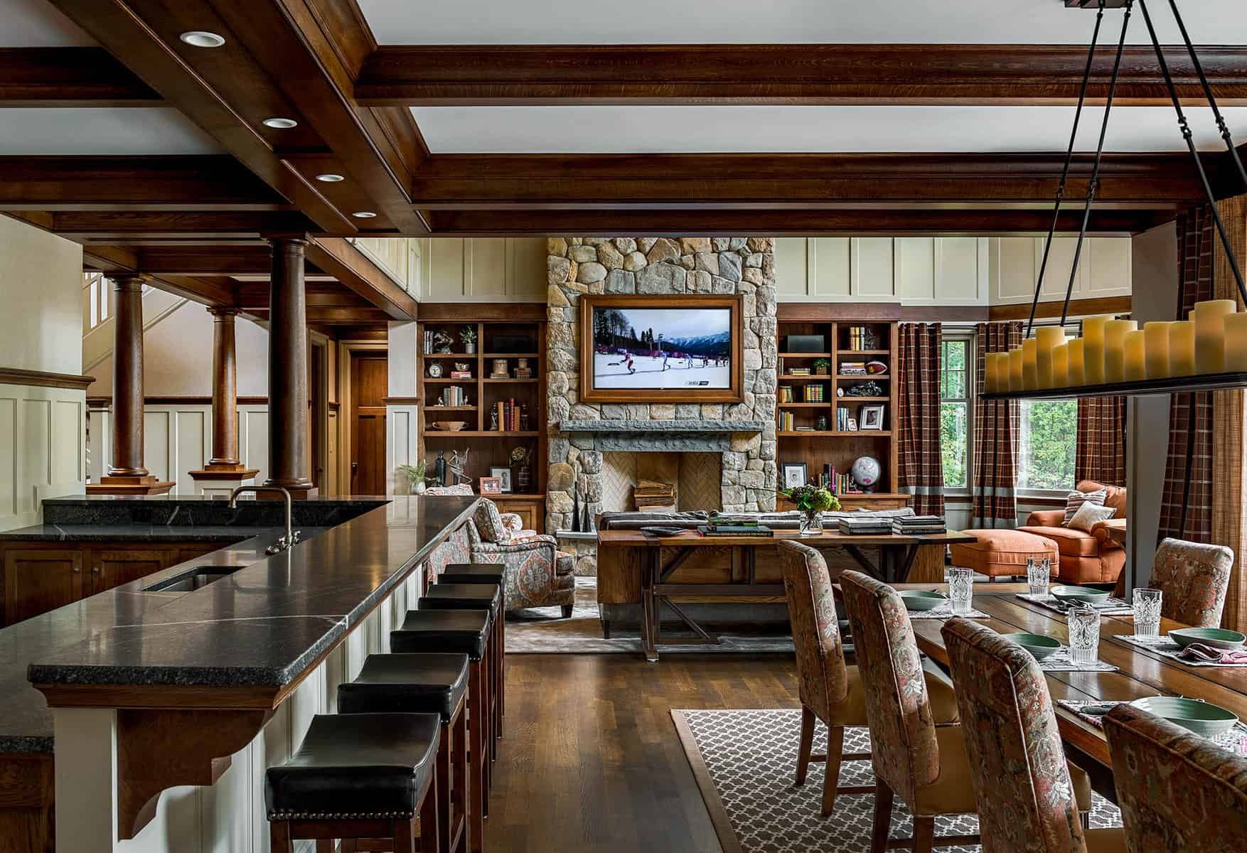 A spacious living room with a large stone fireplace, wooden built-in shelves, a mounted TV, a dining table with chairs, a sofa, and large windows with plaid curtains.