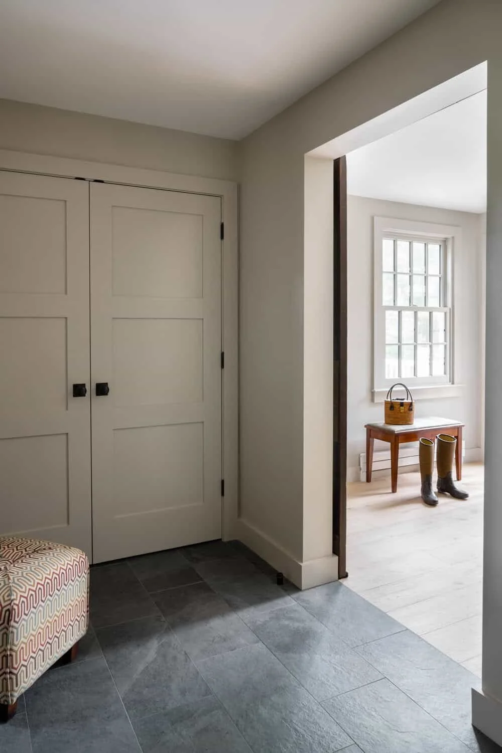 An interior view shows a doorway with closed white double doors and a section of a room with a window, a small wooden table, a woven basket, and a pair of rubber boots on the floor.