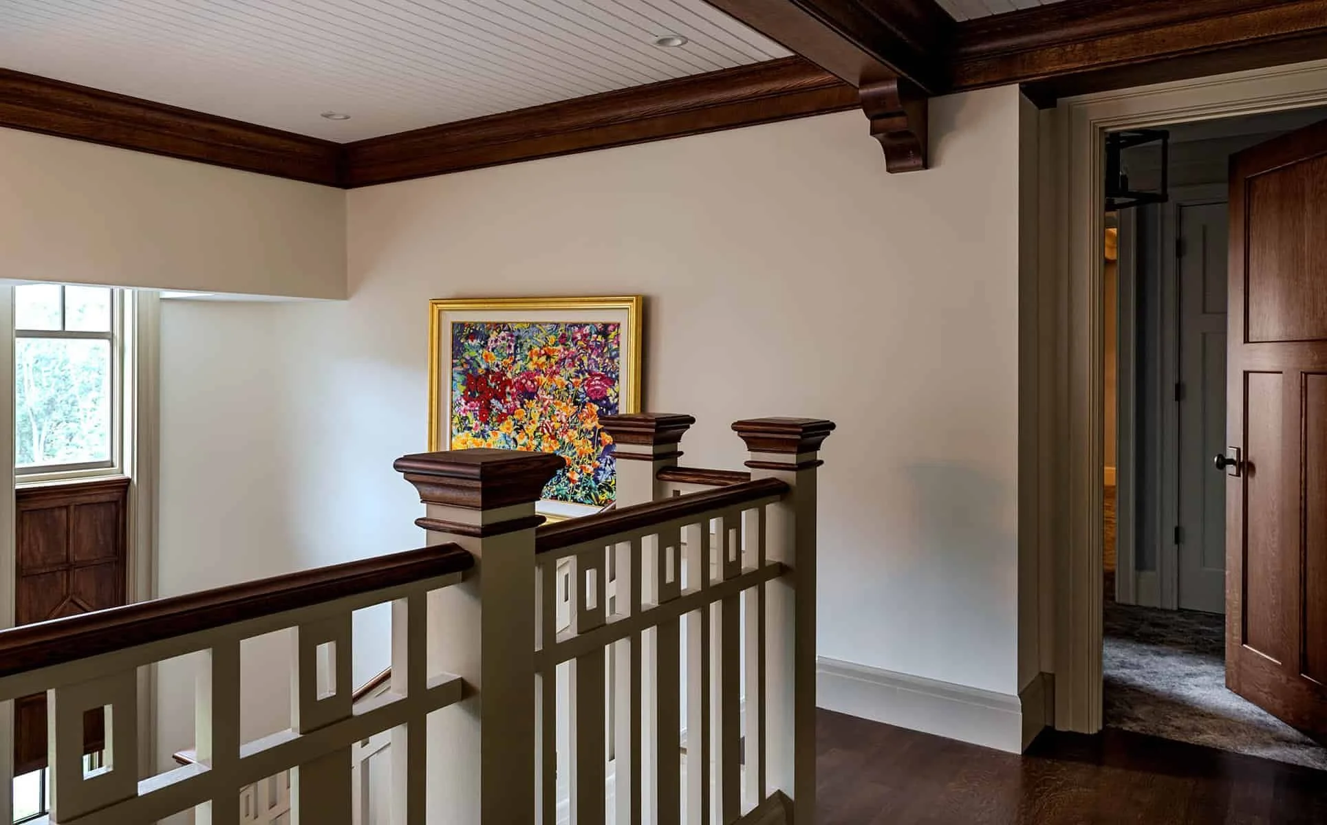 Interior view of a house with wooden trim, a staircase with white banisters, a window on the left, a colorful floral painting on the wall, and an open door leading to a room with carpeted flooring.