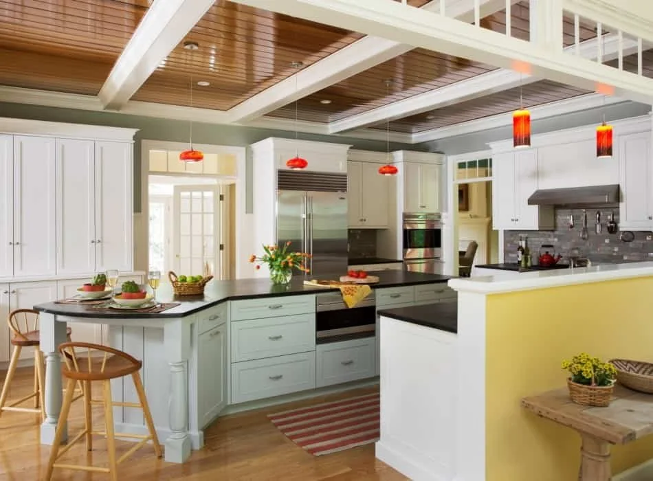 Bright kitchen with white cabinets, a black countertop island, stainless steel appliances, and red pendant lights hanging from a wood-paneled ceiling. Small table with chairs, a flower arrangement, and a yellow accent wall.